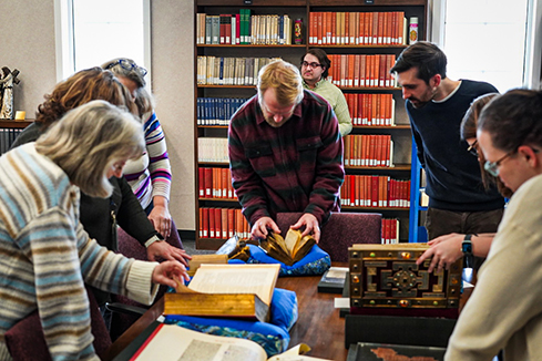 A group of people gather around a collection of ancient texts and books and compare the material to the pages of The Saint John’s Bible. 